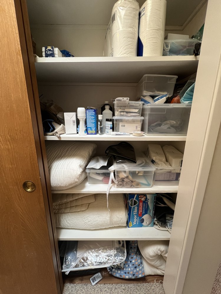 Disorganized bathroom hall closet with toiletries, cleaning supplies, and blankets randomly stored