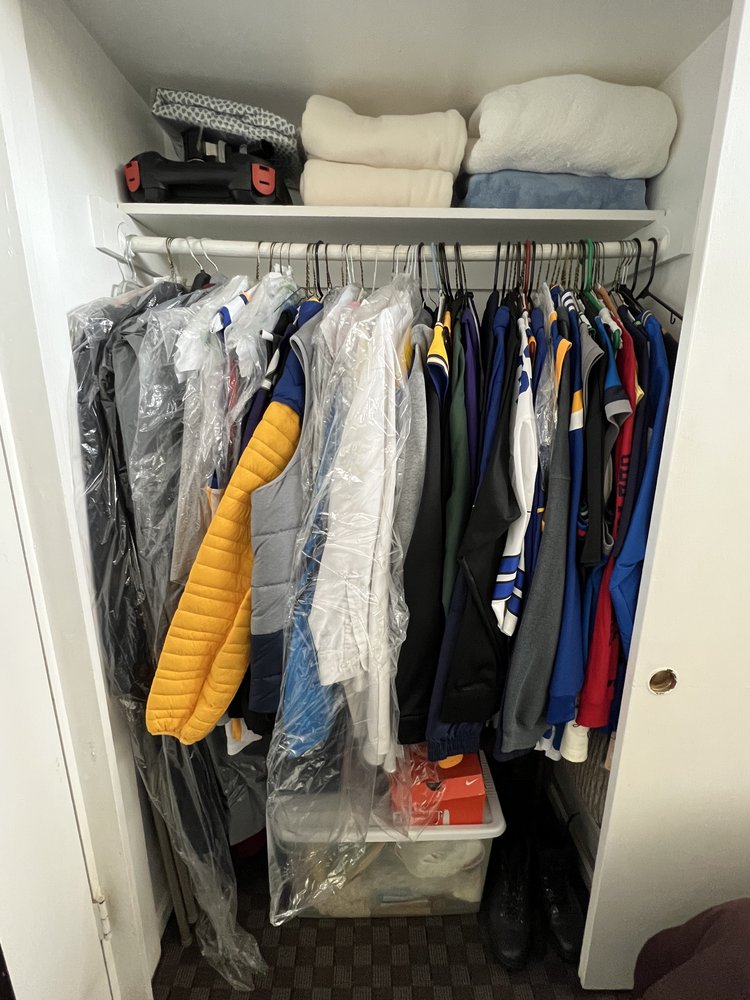 Cluttered reach-in bedroom closet with mixed sports jerseys, jackets, dry-cleaning bags, blankets and accessories on top shelf
